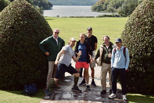 A group of six men are cheerfully posing for a photo on a stone path, flanked by large, manicured topiary bushes